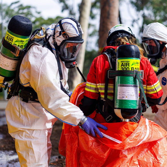 Person in protective gear with air tanks assisting another person in an orange suit outdoors.