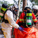 Person in protective gear with air tanks assisting another person in an orange suit outdoors.